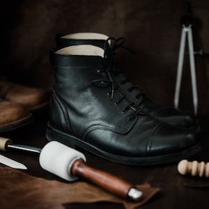May include: Close-up of two pairs of leather boots, one brown and one black, with tools for shoemaking on a brown leather surface.
