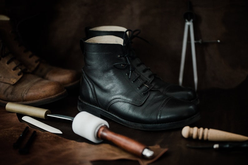 May include: Close-up of two pairs of leather boots, one brown and one black, with tools for shoemaking on a brown leather surface.