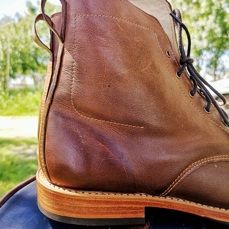 May include: Close-up of a brown leather boot with a lace-up closure and a brown leather sole. The boot has a distressed look and is made of high-quality leather.