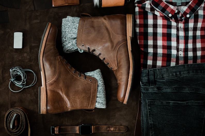 May include: A pair of brown leather boots with grey socks, a red and black plaid shirt, a pair of dark blue jeans, a leather belt, and a lighter.
