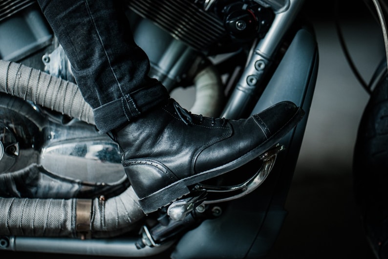 May include: Close-up of a black leather motorcycle boot with a cap toe, worn by a person riding a motorcycle. The boot is resting on the motorcycle's foot peg.