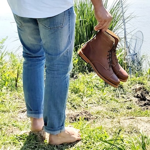 May include: A person wearing blue jeans is holding a pair of brown leather lace-up boots with a cap toe.