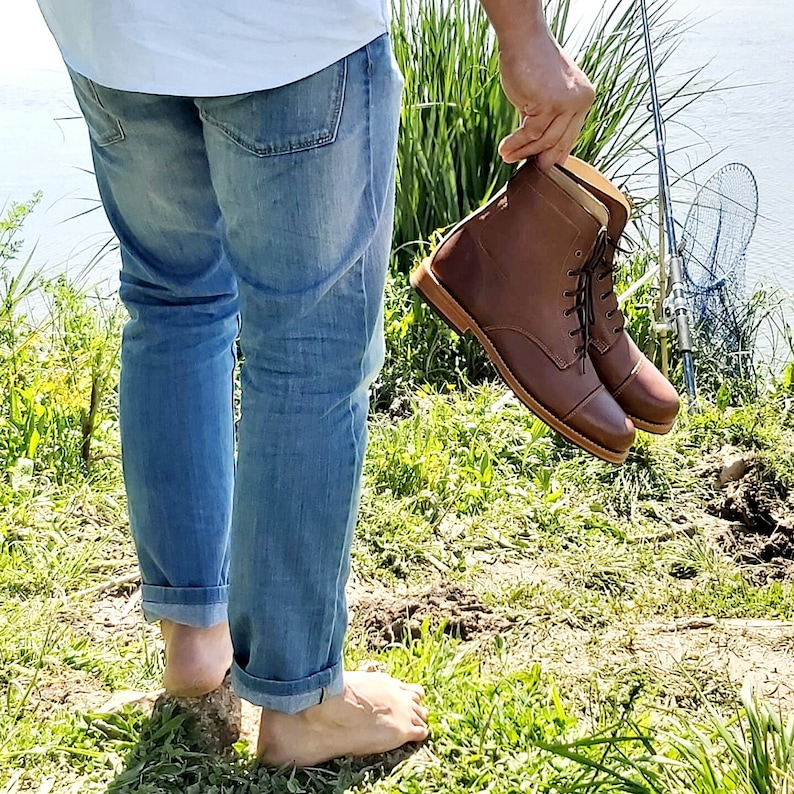 May include: A person wearing blue jeans is holding a pair of brown leather lace-up boots with a cap toe.