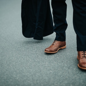 May include: A person wearing dark blue pants and brown leather shoes with laces. The person is walking on a gray asphalt road.