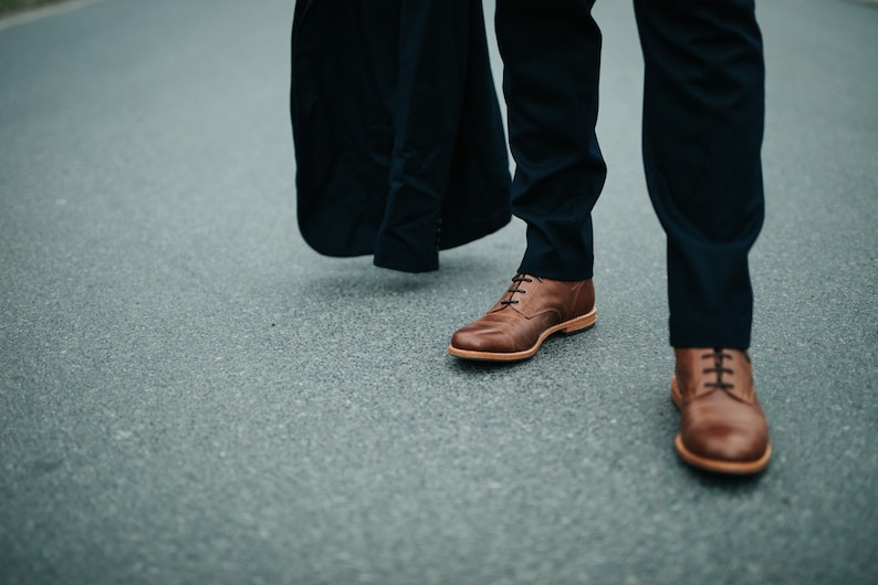 May include: A person wearing dark blue pants and brown leather shoes with laces. The person is walking on a gray asphalt road.