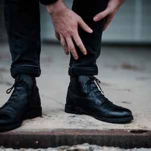 May include: A pair of black leather lace-up boots with a rounded toe. The boots are worn by a person with rolled up black jeans.