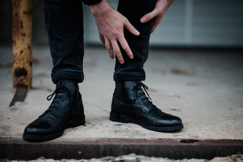 May include: A pair of black leather lace-up boots with a rounded toe. The boots are worn by a person with rolled up black jeans.