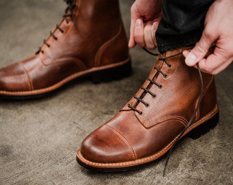 Puede incluir: Un par de botas de cuero marrón con cordones y punta redonda. Las botas tienen una suela gruesa y están atadas con cordones marrones.