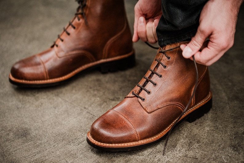 Puede incluir: Un par de botas de cuero marr&oacute;n con cordones y punta redonda. Las botas tienen una suela gruesa y est&aacute;n atadas con cordones marrones.