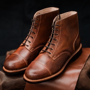 Puede incluir: Un par de botas de cuero marr&oacute;n con cordones y puntera de captoe. Las botas est&aacute;n sobre una superficie de cuero marr&oacute;n.
