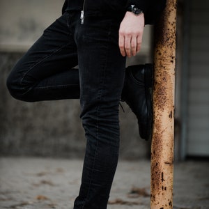 May include: A person wearing black jeans and a black jacket, standing with one leg bent and resting on a rusty metal pole.