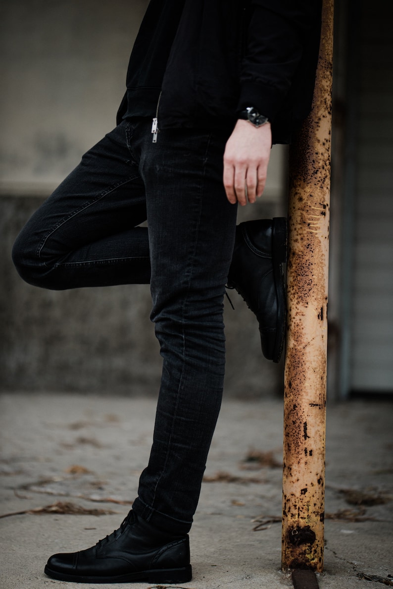 May include: A person wearing black jeans and a black jacket, standing with one leg bent and resting on a rusty metal pole.