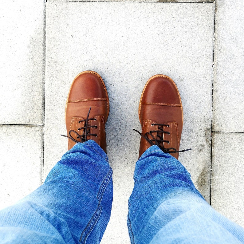May include: A pair of brown leather lace-up boots with a person wearing blue jeans.