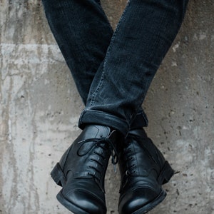 May include: A pair of black leather lace-up boots with a rounded toe. The boots are worn by a person wearing dark blue jeans.