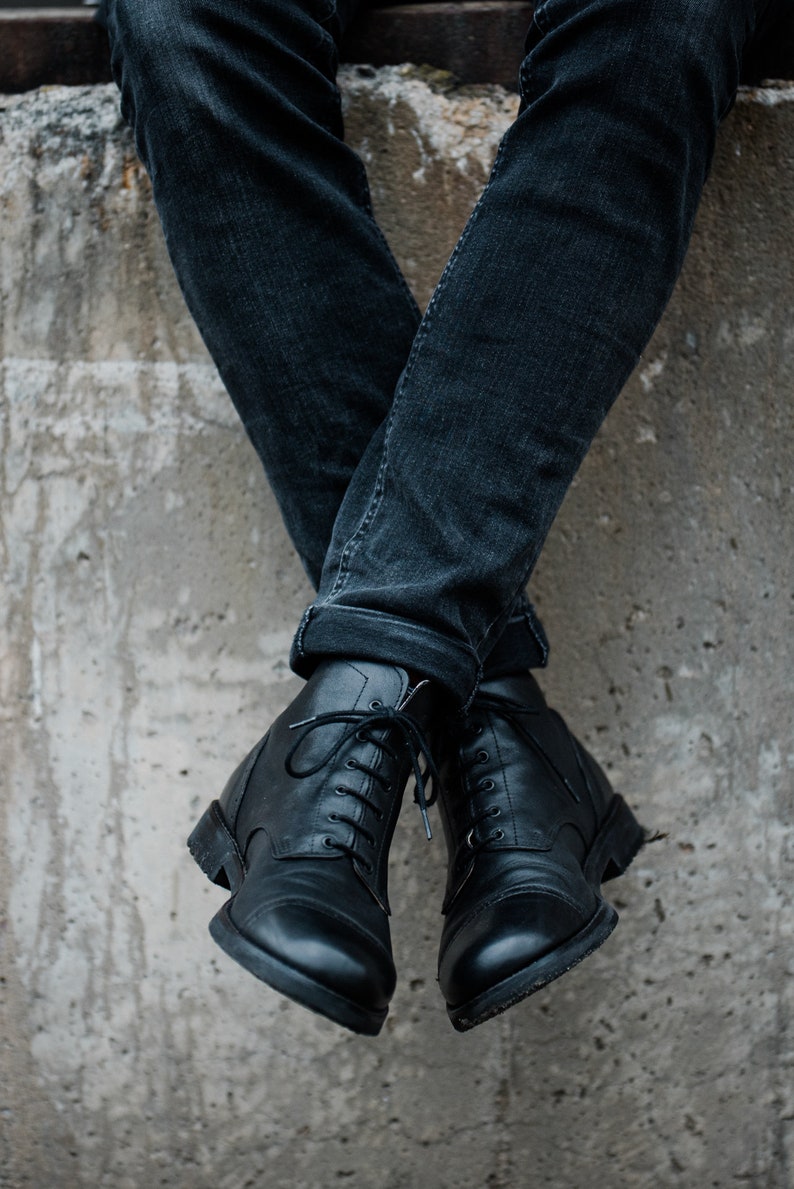 May include: A pair of black leather lace-up boots with a rounded toe. The boots are worn by a person wearing dark blue jeans.