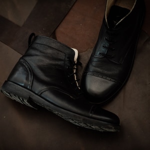May include: Two pairs of black leather boots with laces. One boot is a lace-up ankle boot with a rounded toe. The other boot is a low-top boot with a cap toe.