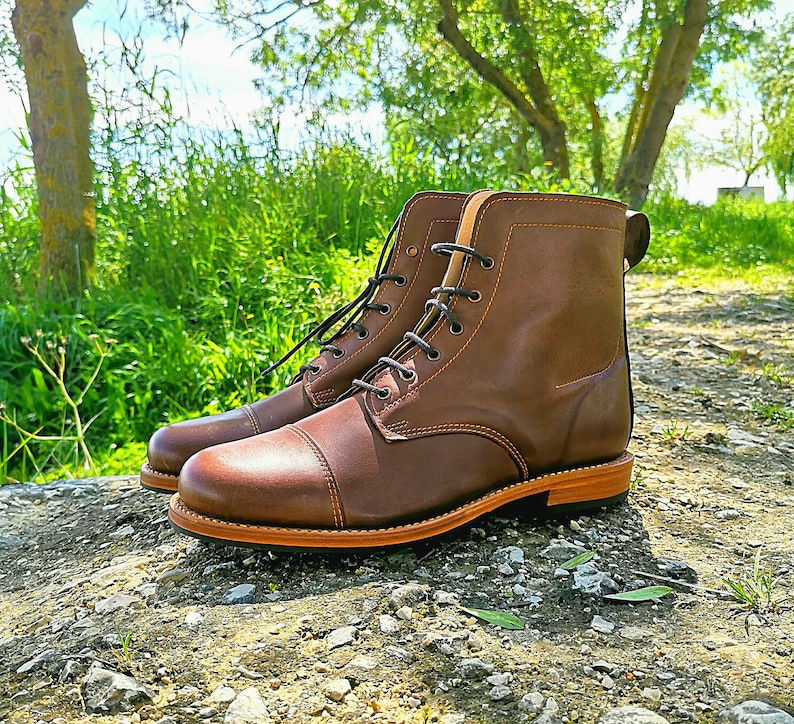 May include: Brown leather lace-up boots with a cap toe design. The boots have a thick sole and are set against a backdrop of green grass and trees.