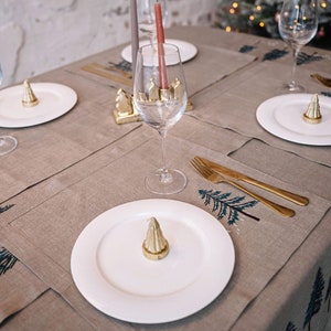 May include: A table setting with a beige linen tablecloth and placemats featuring a blue pine tree design. Each place setting includes a white plate, a gold metal ornament, a gold fork and knife, and a clear wine glass.