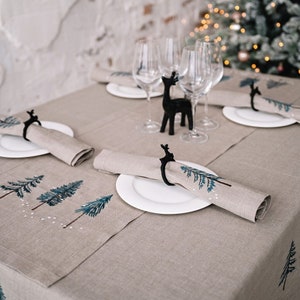 May include: A beige linen tablecloth with a winter forest design. The tablecloth is set with white plates and napkins with black reindeer napkin rings. Two wine glasses are on the table.