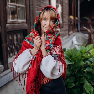 Peut inclure: Une femme portant un foulard traditionnel rouge et blanc brodé avec des motifs floraux. Le foulard est drapé sur ses épaules et noué au cou. Elle porte une blouse blanche avec des détails brodés et une jupe noire.