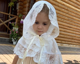 Può includere: Un berretto bianco in pizzo con un delicato motivo floreale. Il berretto ha un cappuccio arricciato e un nastro da legare. Il berretto è indossato da una bambina con i capelli castano chiaro.