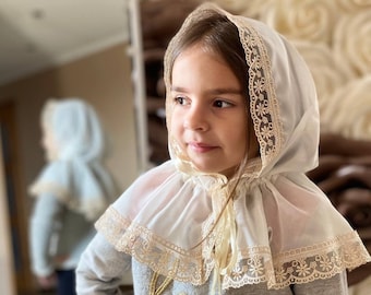 Può includere: Una bambina che indossa un berretto bianco di pizzo con un colletto a balze. Il berretto ha un delicato motivo floreale ed è legato con un nastro sotto il mento. Indossa un maglione grigio con ricami dorati.