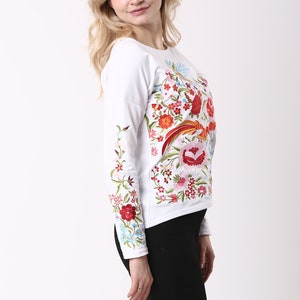 May include: A white long-sleeved top featuring colourful floral embroidery in red, orange, pink, and green. The top is worn with black trousers and black high-heeled shoes. The woman is smiling and posing.