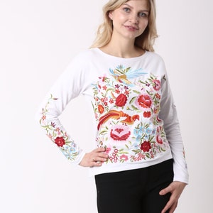 May include: White long-sleeved shirt with vibrant floral and bird embroidery. The design includes red, pink, blue, and green flowers and birds. The embroidery extends to the sleeves. The shirt is worn with black trousers.