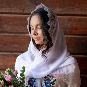 May include: A white head covering featuring intricate lace trim along the edges, worn over a light-colored garment with colorful floral embroidery. The head covering is made of a lightweight, semi-sheer fabric that drapes softly around the head.