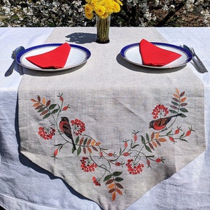 May include: A white tablecloth with a beige table runner featuring an embroidered design of red berries and birds. Two white plates with red napkins are set on the table. A vase with yellow flowers sits in the centre of the table runner.