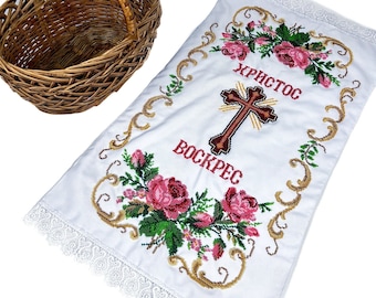 May include: A white embroidered cloth with a cross and the text "ХРИСТОС ВОСКРЕС" in Cyrillic script. The cloth has a lace border and is decorated with pink roses. A brown wicker basket is also in the image.