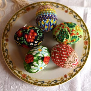 Puede incluir: Seis huevos de Pascua decorativos, cada uno intrincadamente adornado con patrones coloridos de cuentas. Los huevos descansan sobre un plato decorativo con un borde floral. Los diseños incluyen motivos florales, geométricos e inspirados en la naturaleza.