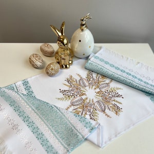 Puede incluir: Un camino de mesa blanco con un borde bordado verde y blanco. El centro del camino de mesa presenta un diseño bordado de una corona de huevos marrones y dorados con hojas verdes. Hay cuatro huevos pintados en el camino de mesa, dos de los cuales son marrones y dos son blancos. También hay dos figuritas de conejito doradas en el camino de mesa.