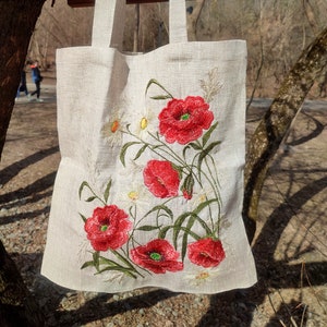 Könnte beinhalten: Eine weiße Leinentasche mit gestickten roten Mohnblumen und weißen Gänseblümchen. Die Tasche hat zwei lange Henkel.