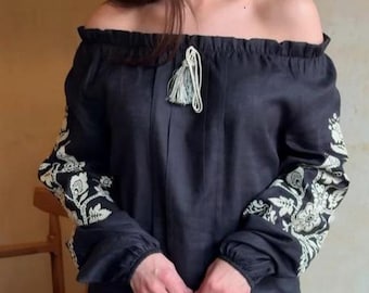 May include: A black off-the-shoulder blouse with long sleeves. The sleeves feature white floral embroidery. The blouse has a tassel detail at the neckline. The fabric appears to be linen or a similar lightweight material.
