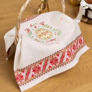 Op de afbeelding: Een gevlochten mand met een wit geborduurde doek met een bloemmotief en rode accenten. Een gouden konijnenbeeldje en een gouden ei staan op de houten tafel. Een gebakken brood met een lint staat ook op de tafel.