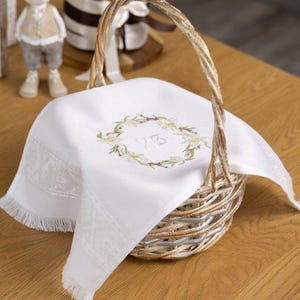 Peut inclure: Un panier en osier avec un tissu blanc brodé d'une couronne florale. Le panier est posé sur une table en bois, avec un lapin décoratif et un vase de tulipes jaunes. L'ensemble évoque Pâques.