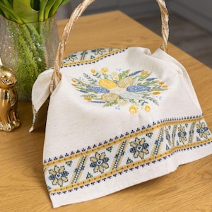 Op de afbeelding: Een decoratieve Paasscène met een gouden ei en konijn, een mand met een bloemen geborduurde doek en een vaas met tulpen met paarse en gele bloemen. De mand heeft een gevlochten handvat en de doek heeft een blauw en geel bloemenpatroon.