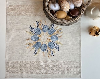 May include: A beige linen table runner with an embroidered wreath of blue and yellow eggs. A wicker basket filled with decorated Easter eggs sits on the table runner.