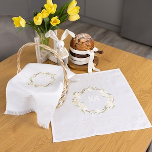 May include: An Easter still life on a wooden table. White linen towels with floral wreath embroidery and the letters "XB" are visible. A basket holds one towel. A vase of yellow tulips, a bunny figurine, and a decorated cake complete the scene.