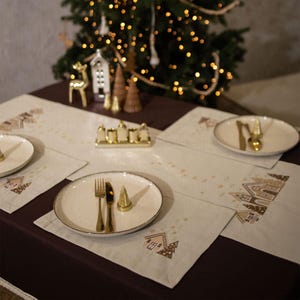 May include: Four white placemats with a gingerbread house design and gold accents. The placemats are set on a brown tablecloth. There are two plates, two forks, two knives, and two gold candle holders on the table.