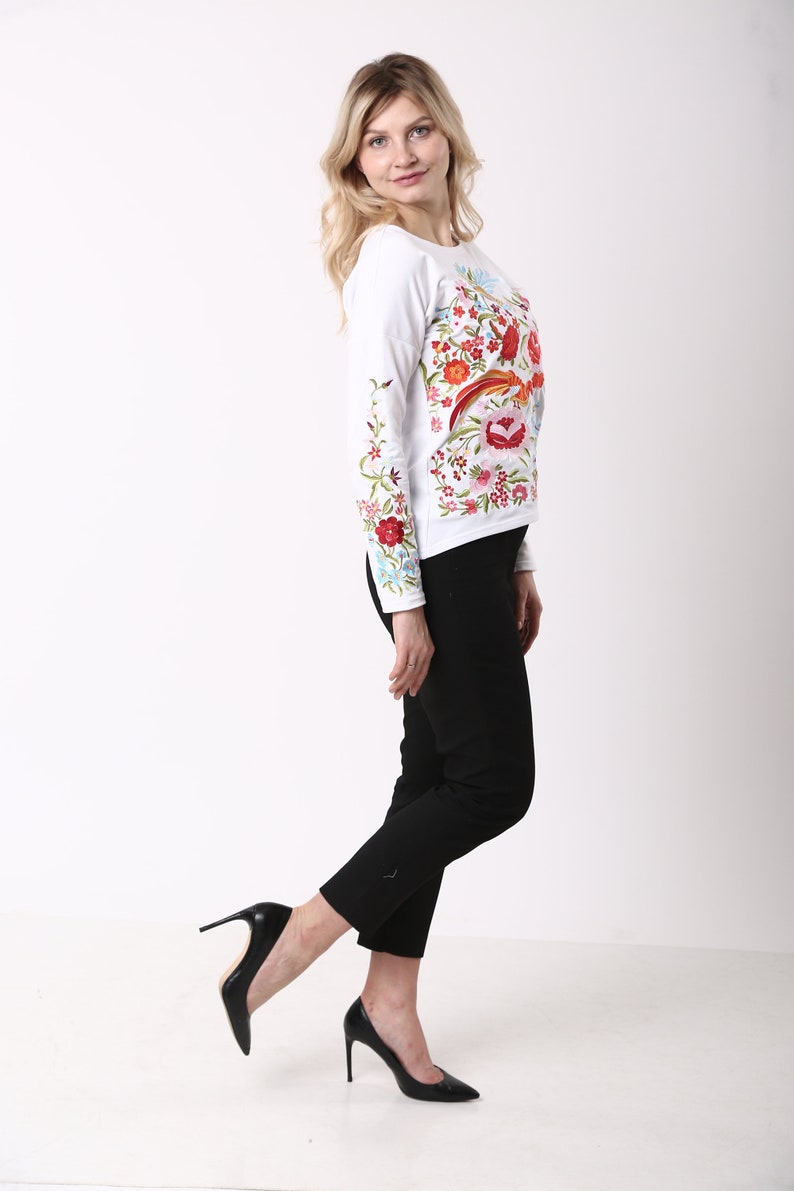 May include: A white long-sleeved top featuring colourful floral embroidery in red, orange, pink, and green. The top is worn with black trousers and black high-heeled shoes. The woman is smiling and posing.