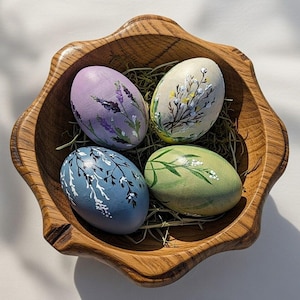 May include: A wooden bowl contains four painted Easter eggs on a bed of straw. The eggs are decorated in pastel shades of lavender, blue, green, and cream, each with a floral pattern. The bowl is made of light brown wood.