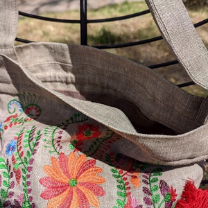 May include: A beige linen tote bag with a red tassel and embroidered floral design featuring red, orange, green, and purple flowers.