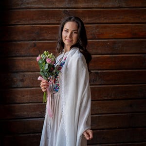 Puede incluir: Una mujer con un chal blanco y un vestido largo y claro con bordados florales. Sostiene un ramo de flores rosas atado con una cinta rosa. El fondo es una pared de madera.