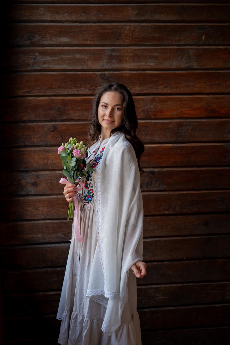 Puede incluir: Una mujer con un chal blanco y un vestido largo y claro con bordados florales. Sostiene un ramo de flores rosas atado con una cinta rosa. El fondo es una pared de madera.