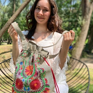 May include: A light gray linen tote bag with colorful embroidered flowers. The bag has red drawstrings with tassels.