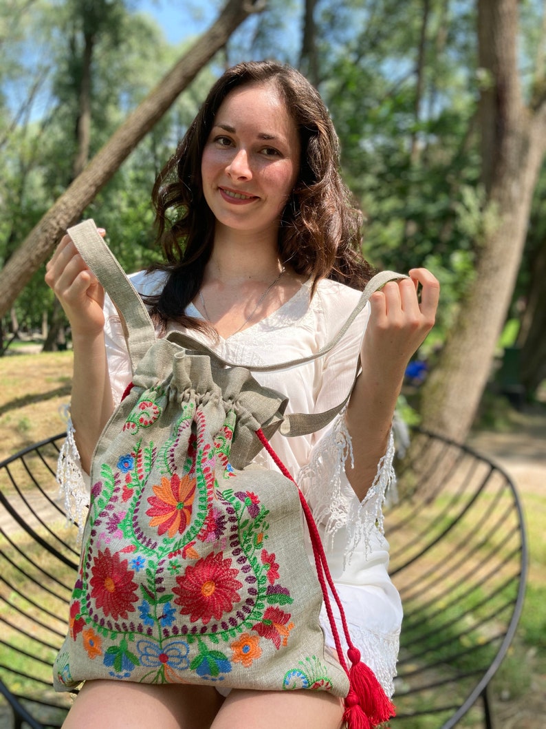 May include: A light gray linen tote bag with colorful embroidered flowers. The bag has red drawstrings with tassels.