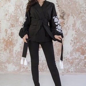 K&ouml;nnte beinhalten: Eine schwarze Wickelbluse mit langen &Auml;rmeln und wei&szlig;en gestickten Blumendesigns an den Manschetten. Die Bluse hat einen Bindeg&uuml;rtel und wei&szlig;e Quasten.
