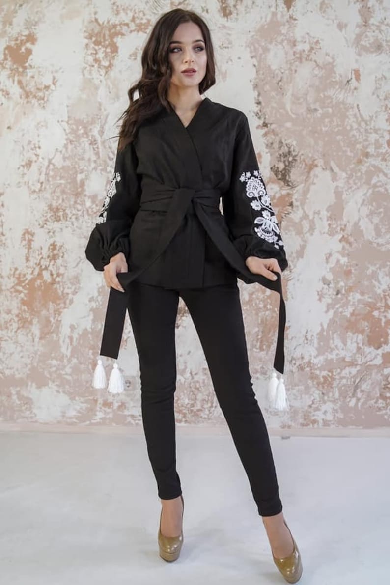 K&ouml;nnte beinhalten: Eine schwarze Wickelbluse mit langen &Auml;rmeln und wei&szlig;en gestickten Blumendesigns an den Manschetten. Die Bluse hat einen Bindeg&uuml;rtel und wei&szlig;e Quasten.